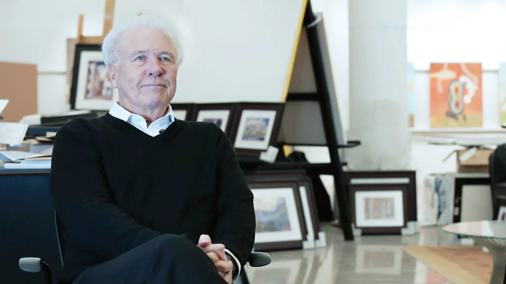 George Acock, an older white man with pure white hair, sits with hands clasped and legs crossed, looking happy, confident and comfortable in a studio where canvasses line the wall and other available space.
