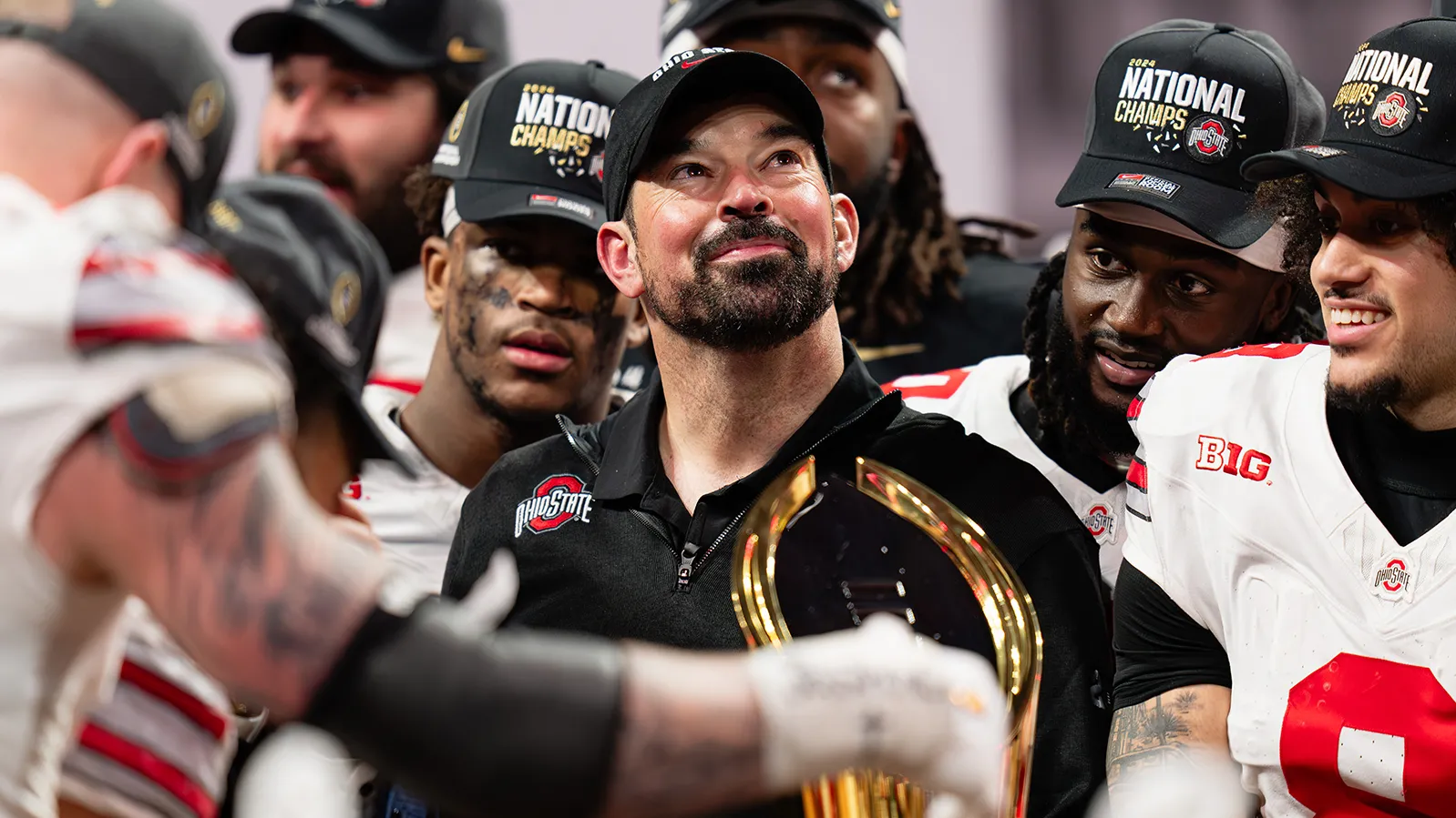 Ohio State football coach Ryan Day, surrounded by football players, looks proudly upward. He appears a little tearful but is smiling. Everyone in the photo taken after they won their final game of the playoffs wears a National Champs baseball cap.