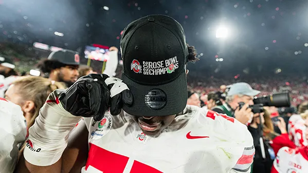 After the bowl game, a young Black player stands amid the crowd of players, media and other on the field and tips his baseball cap down to show the words: 2025 ROSE BOWL CHAMPIONS. The bill of the cap covers all of his face except his lips.