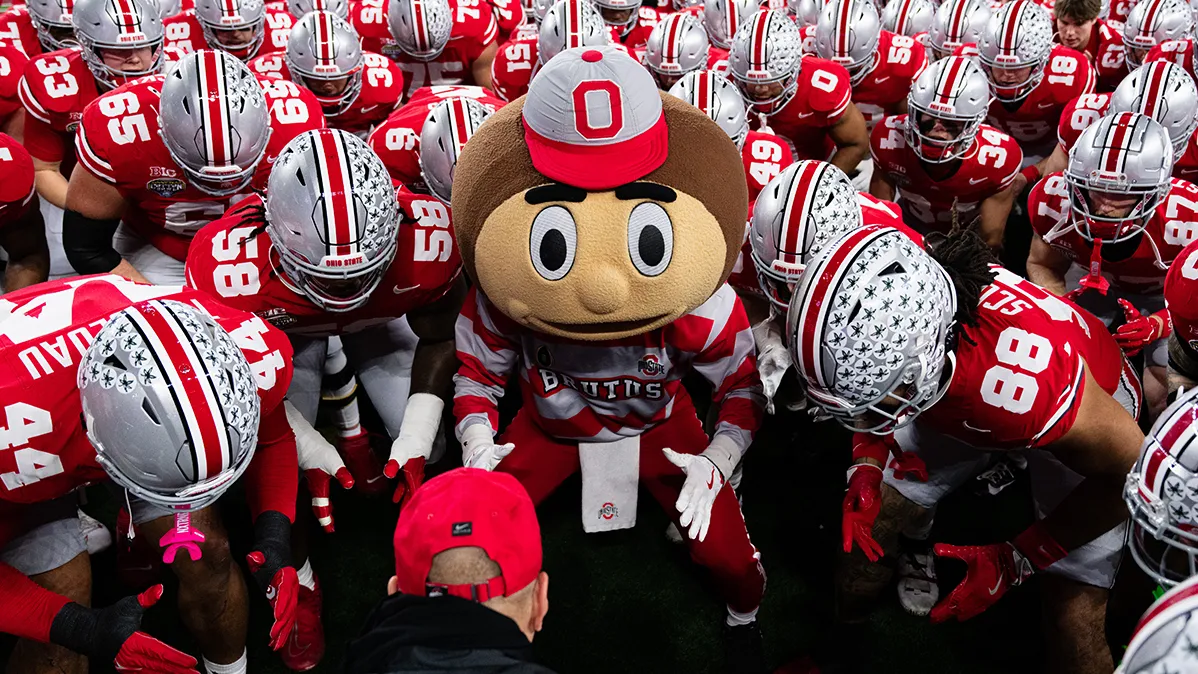 Brutus Buckeye's face is the only one that can be seen as he stands in a huddle of the football team. Everyone else is wearing their helmet and looking down. There are at least 30 players surrounding him