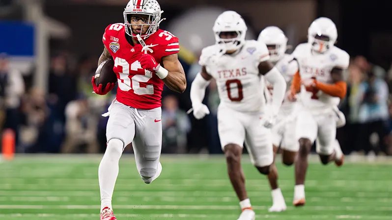 A Buckeye player runs with the football tucked into his right elbow. Three Texas players trail him, and they're not close as they chase him. The Buckeye is a black man with black tape under his eyes and his mouth guard hanging by a short strap from his helmet. He's focused on the end zone.