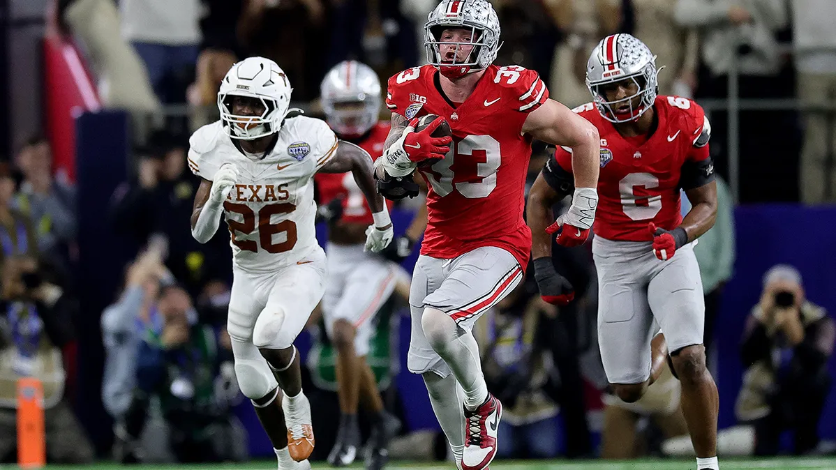 A white football player is red in the face as he runs for all he's worth up the football field. He's a defenseman who picked up the football and will score on this run. One of Texas' offensive players chases and another Buckeye player is running behind him.