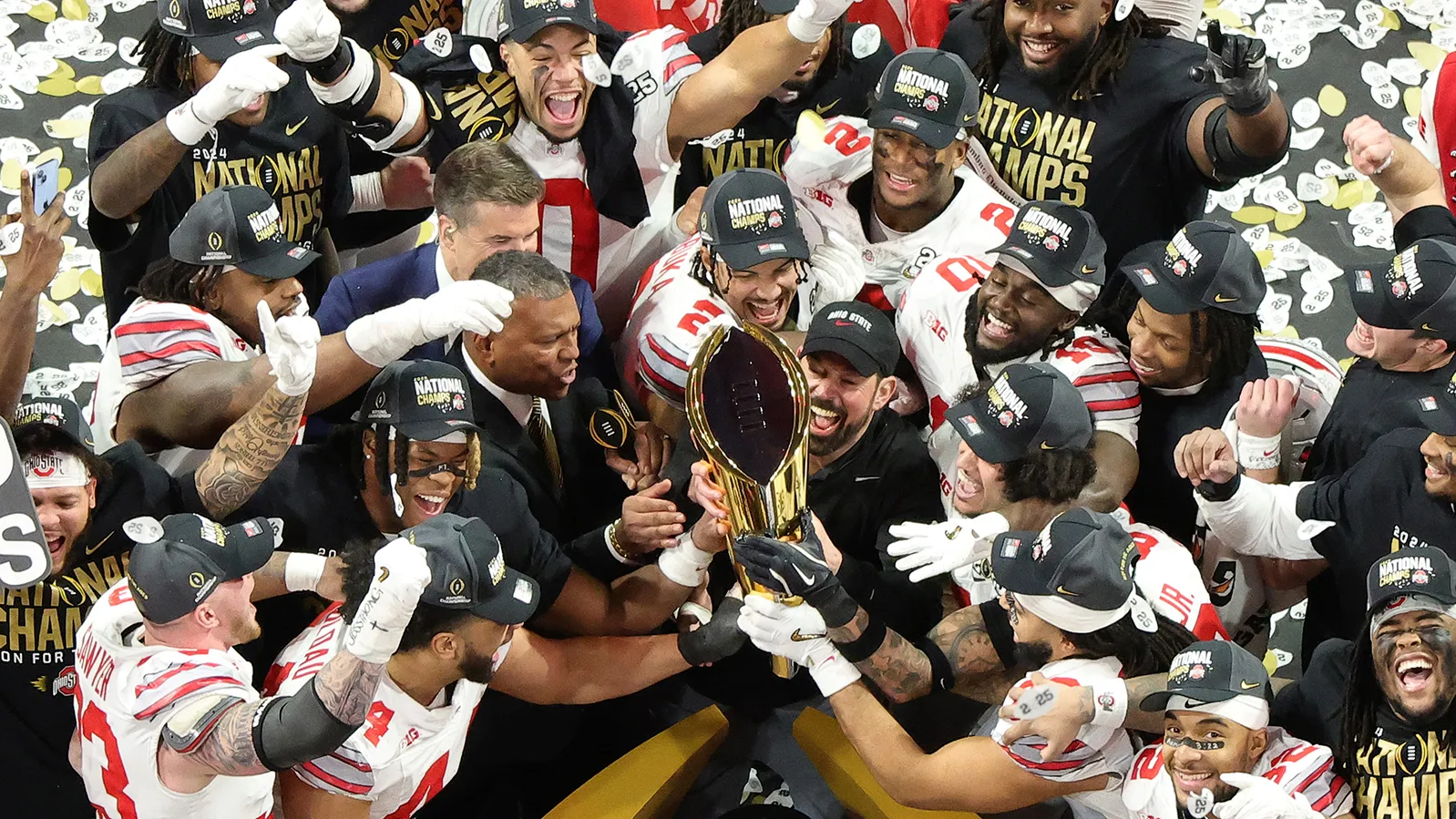 Ryan Day, coaches and players crowd together as they jointly hoist the golden championship trophy. They all wear champion-declaring ballcaps and have gigantic grins in this photo taken from overhead.