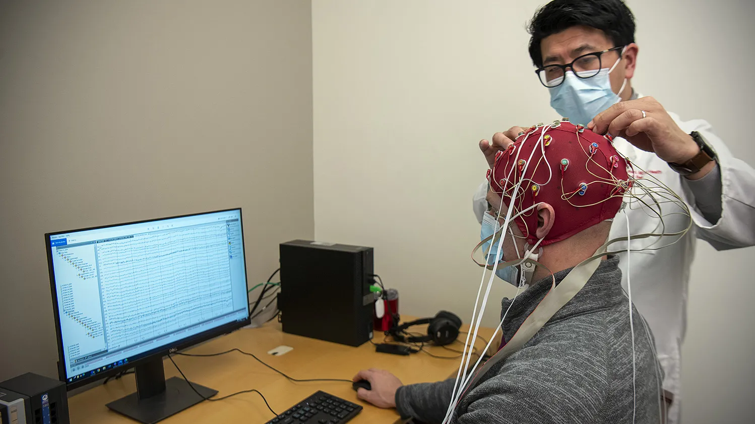 Dr. K. Luan Phan hooks up a stretchy cap with brain monitors to a patient reading a computer monitor. Dr. Phan is an Asian man with glasses and a lab coat. The patient is an older white man. Both wear paper masks.