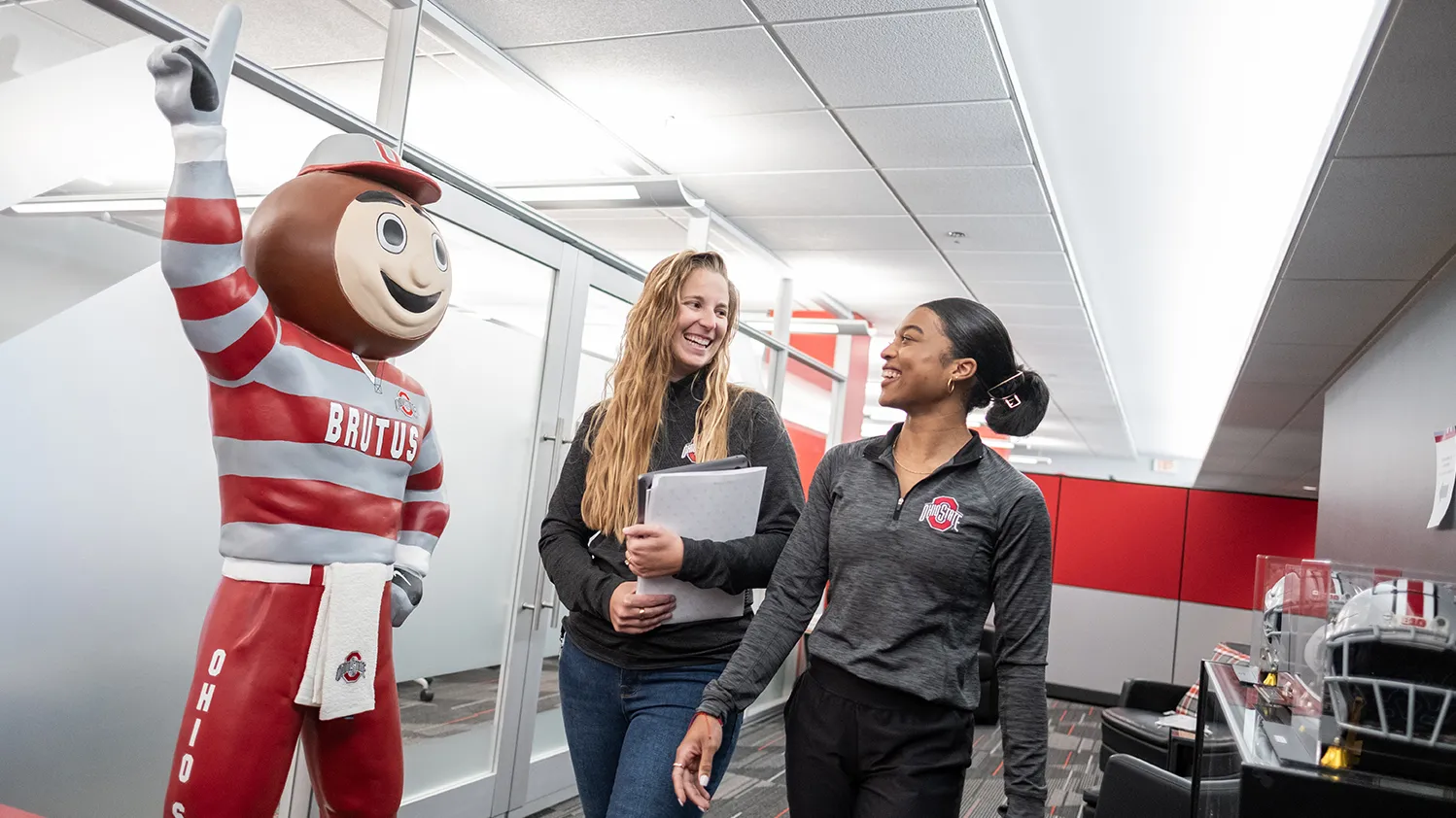 A statue of Brutus Buckeye waves his No. 1 finger as a young white woman and a young black woman walk by happily chatting.