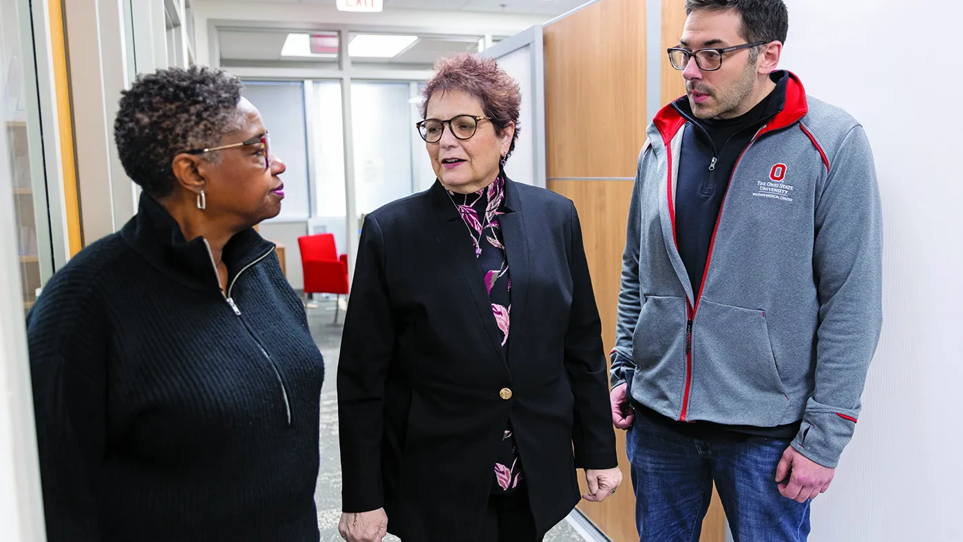 Pausing in an Ohio State building hallway, Electra Paskett speaks with two colleagues. She's a white woman with short hair, glasses and a stylish suit; to her right is a Black woman with short hair and to Electra's left is a tall white man in an Ohio State fleece sweatshirt. They seem invested in their conversation.