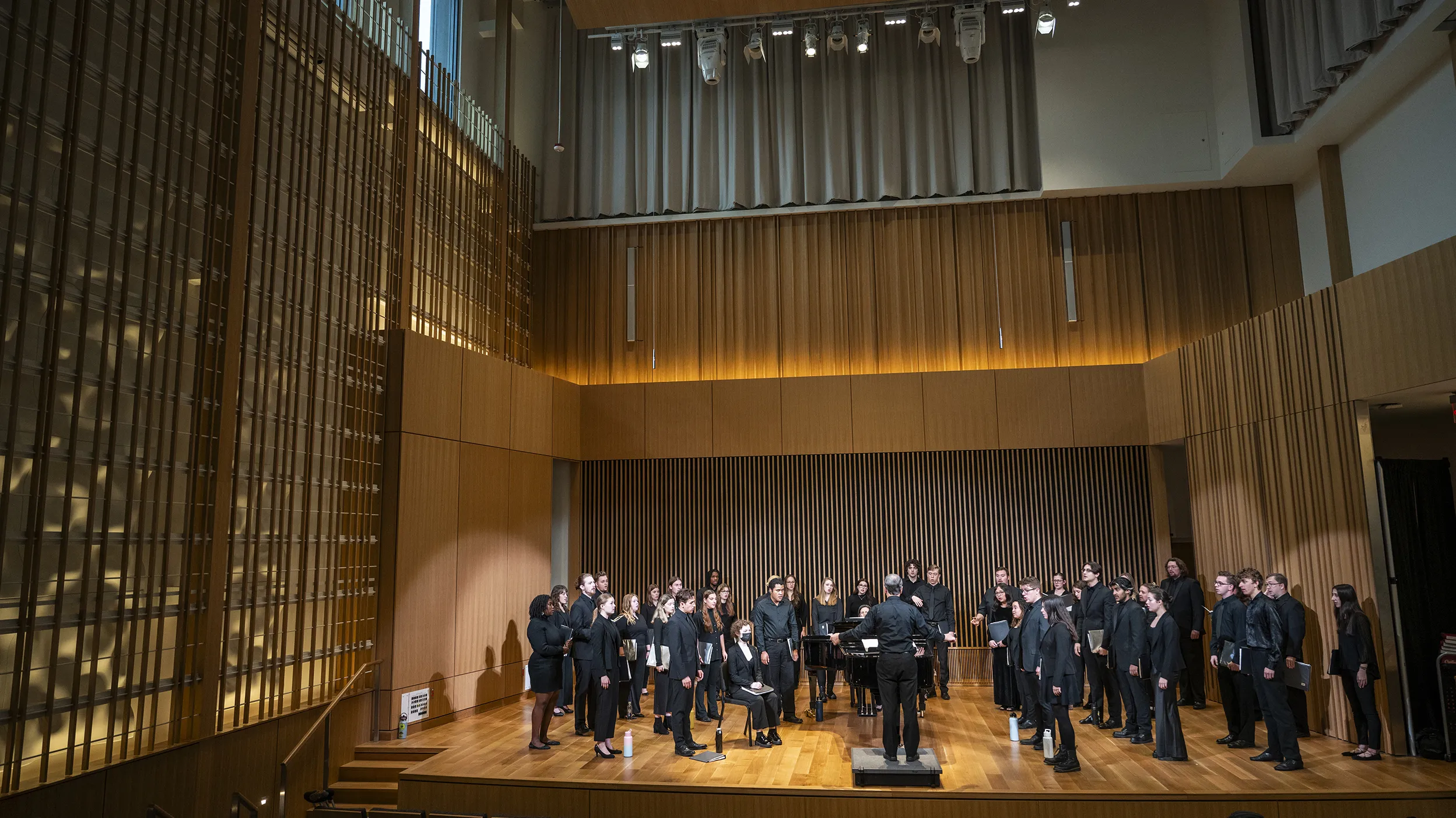 Students standing in rows wearing all black performance outfits singe on a wooden stage as a conductor holds his hands wide. The stage is wrapped in sound optimization and modern-looking elements. The lighting is very warm.
