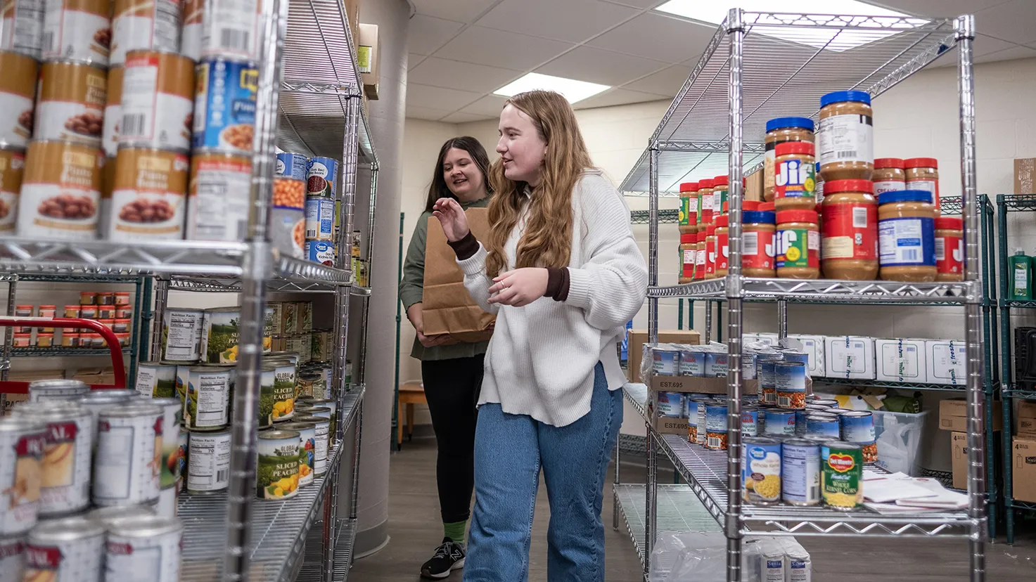 Two student workers, young white women in casual clothes, choose goods from shelves at the student food pantry.