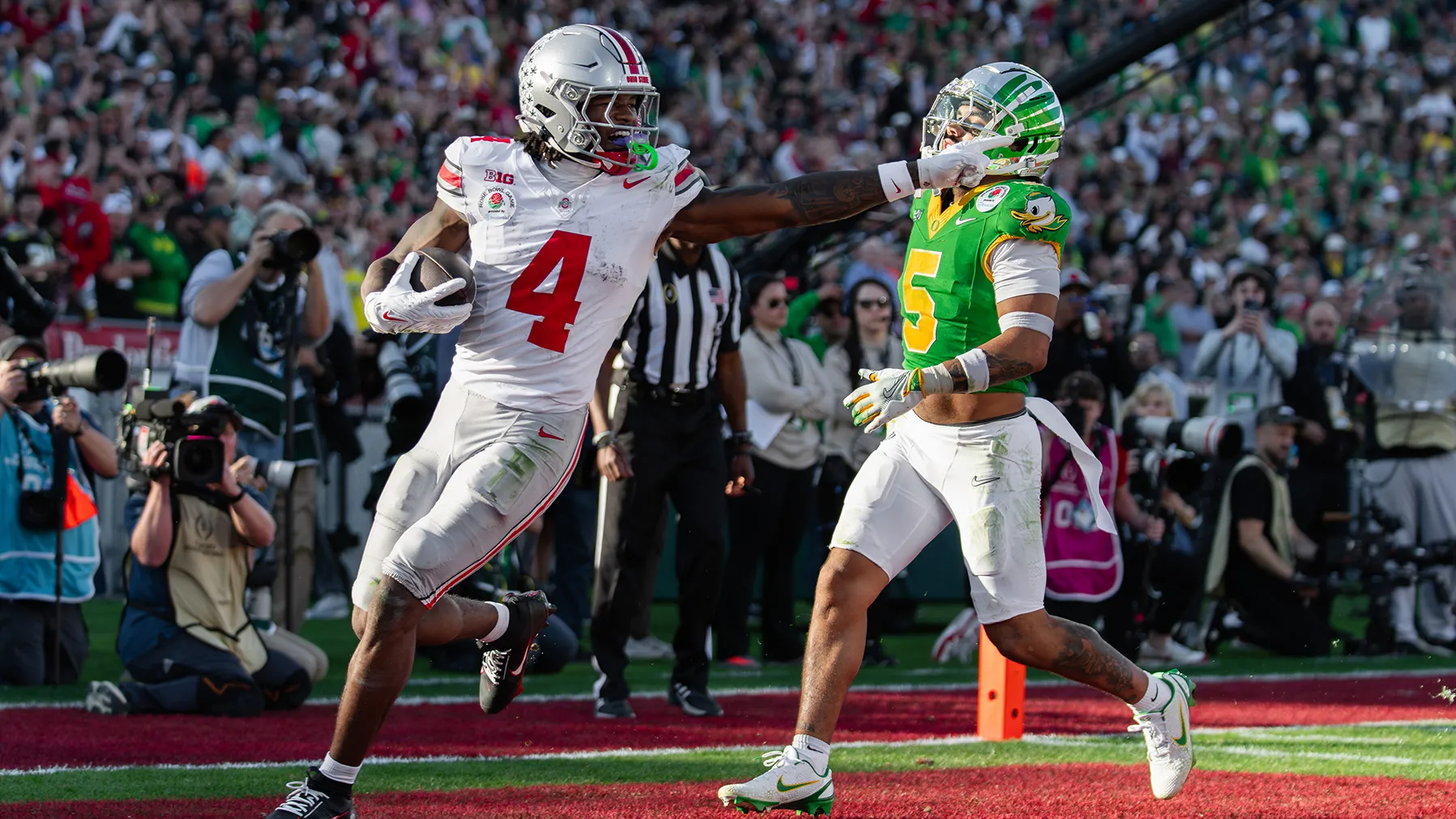 A Buckeye football player, a young Black man with braids peaking out from his helmet, points toward the field right after he runs across the goal line to score a touchdown. The opposing player also slowing down behind him, looks dejected.