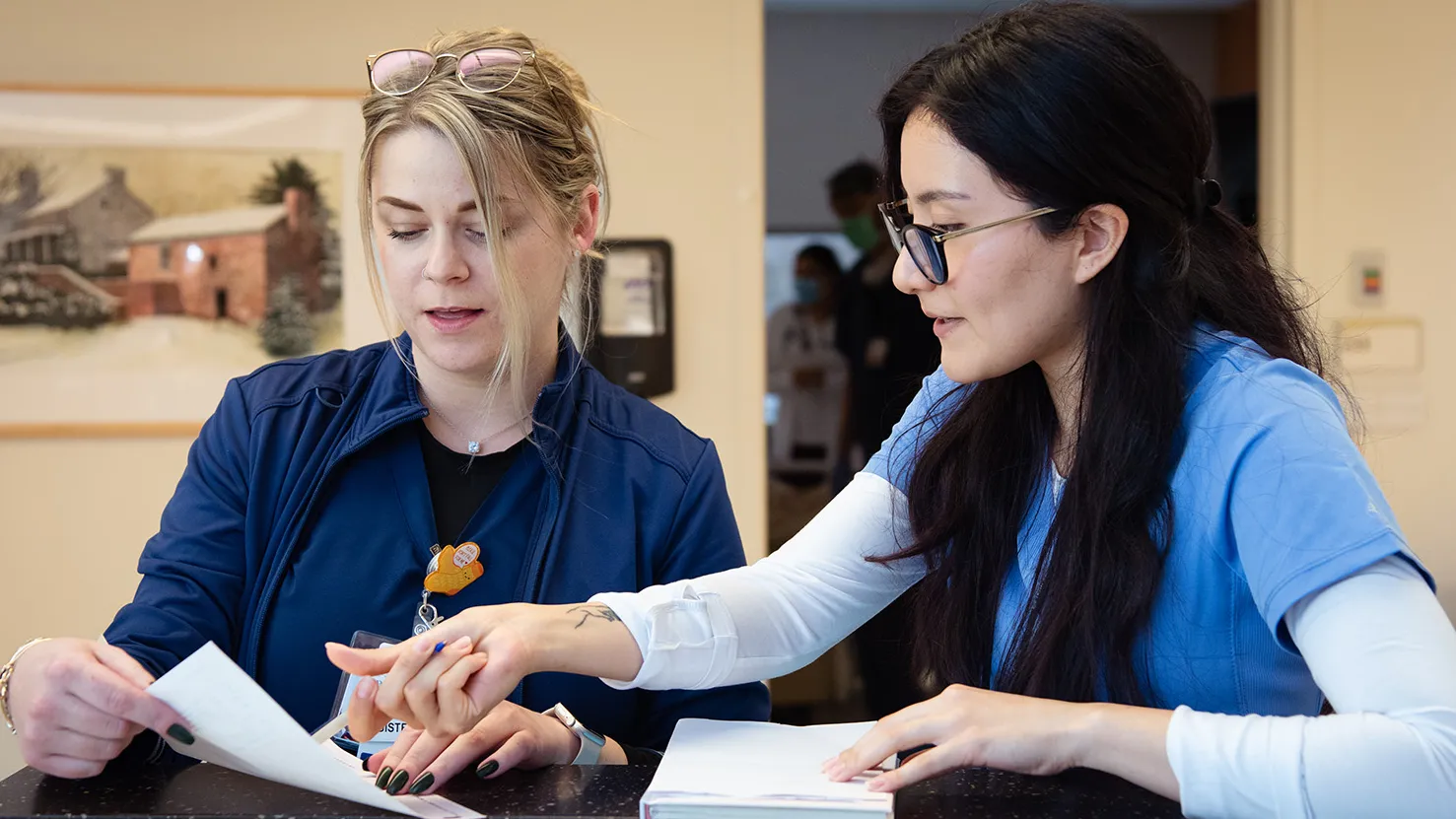 A young woman wearing scrubs works in a medical office. She stands at a counter and points something out on a piece of paper held by another woman. The grad has long dark hair, glasses and a focused expression.