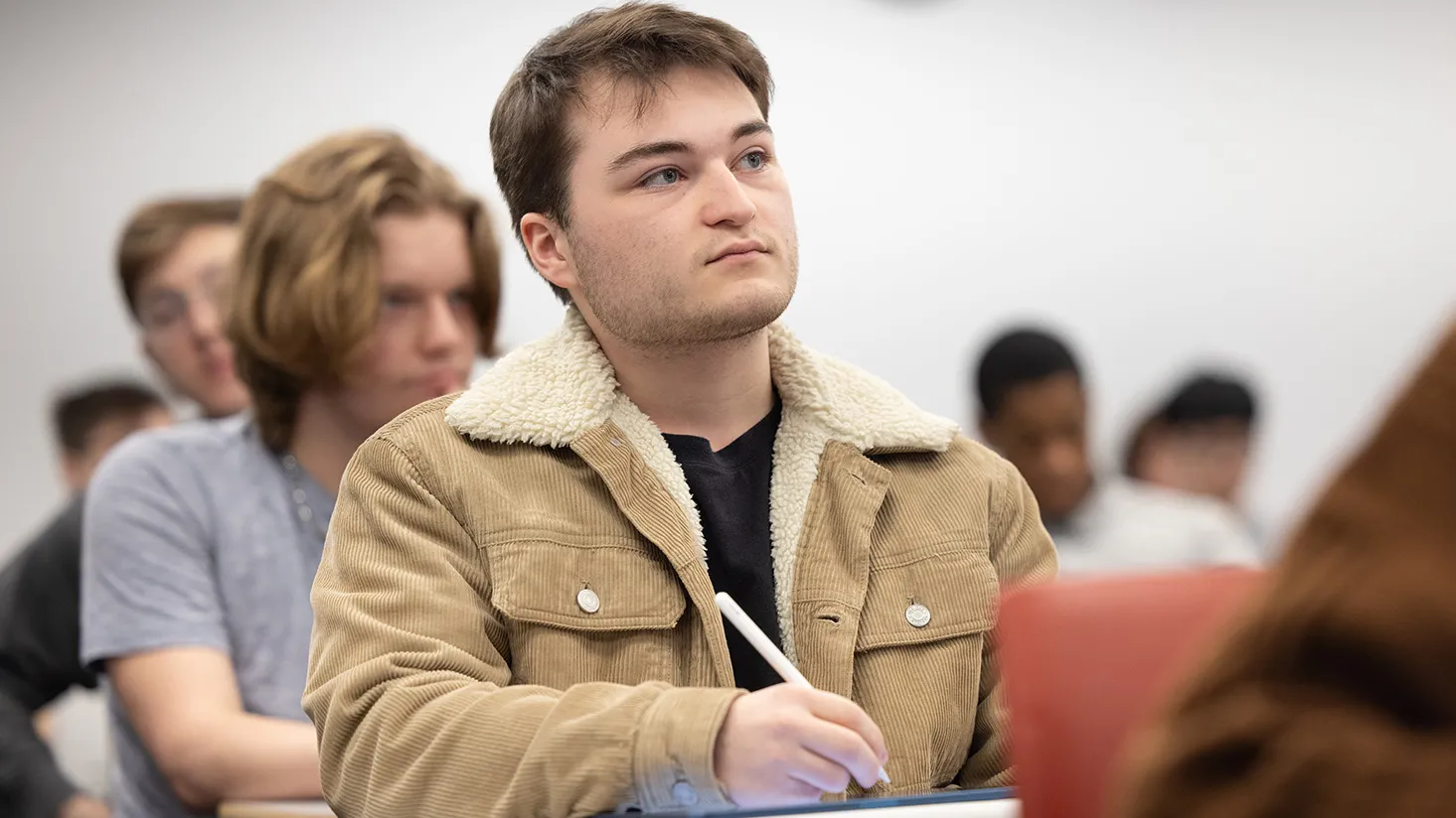A young white man sitting in a filled college classroom intently listens to his professor (who's not in the photo) and takes notes