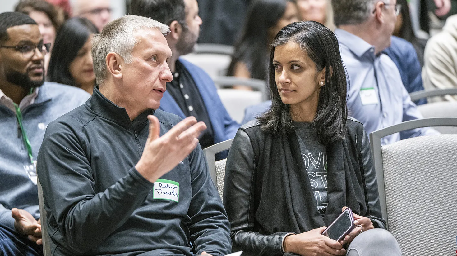 Ratmir Timashev, a white man with short white hair, gestures as he sits at an event and speaks to the woman beside him, Shereen Agrawal. She is a petite woman on Indian descent wearing a chic jacket over her T-shirt and Jeans. They're discussion seems serious, and many people of all races and genders fill seats behind their row.