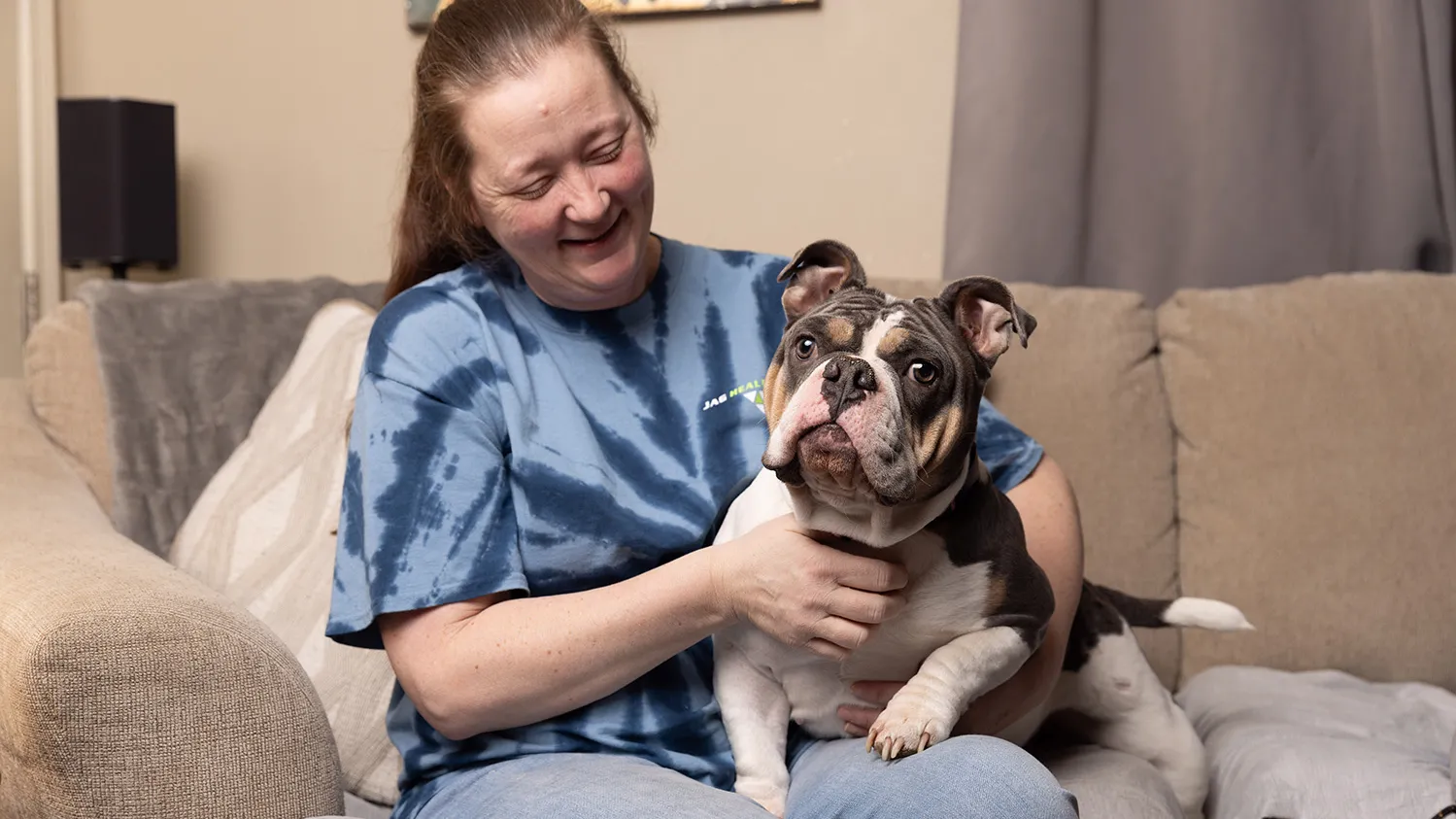 A woman smiles as she looks down at the dog she's holding on her lap. Her pet is an English bulldog with charming face wrinkles and multicolored spots. It is slightly bug eyed. All together it looks like it'd enjoy some pets from the viewer.