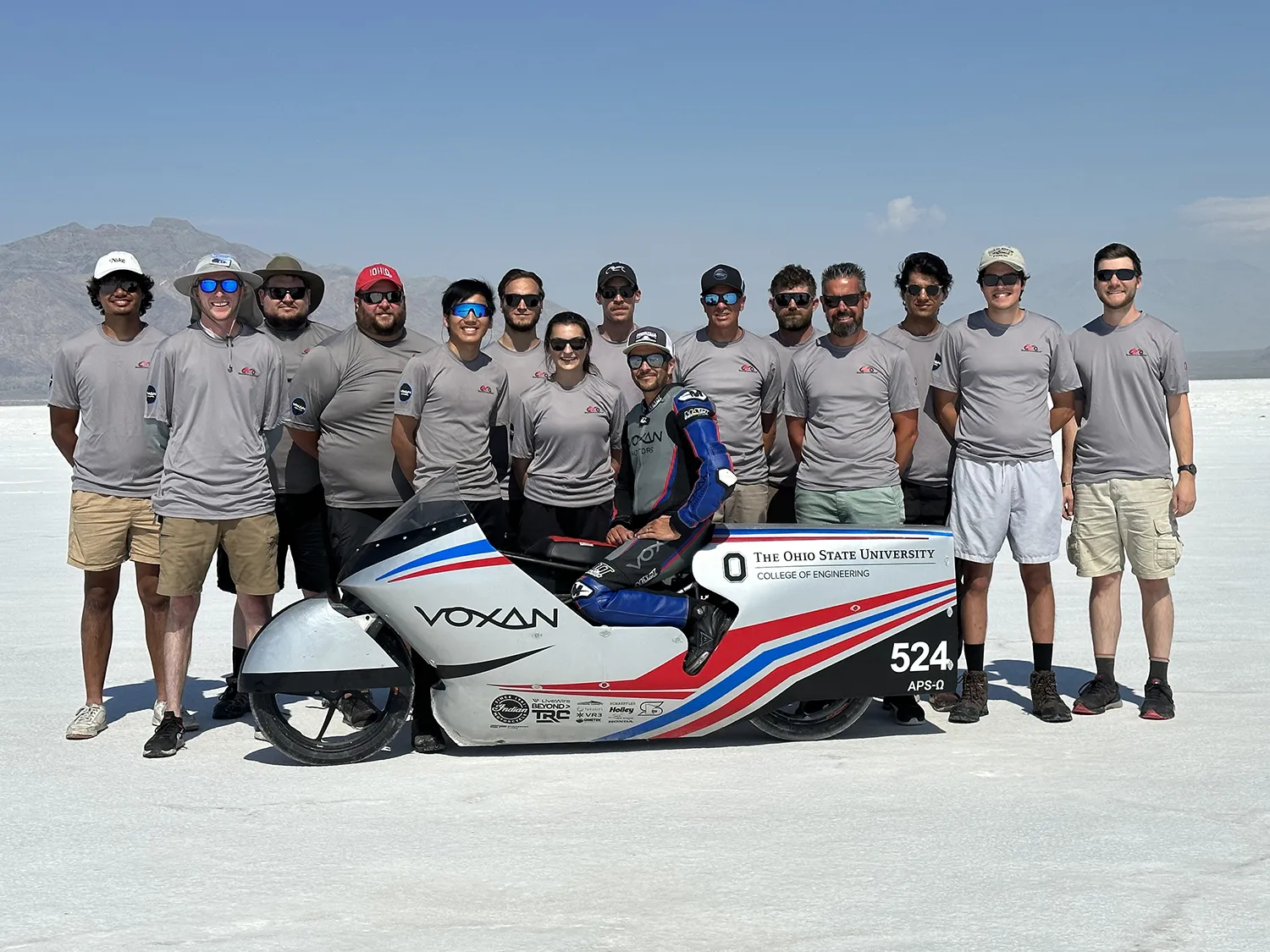 The Ohio State electric motorcycle team poses with their record breaking creation in the Bonneville Saltflats in northwestern Utah. The salt flats see like a beach, but there is no water and the sand is hard. In teh background are mountains. The team is 13 men plus Laura Friedmann and the driver. They all pose proudly.