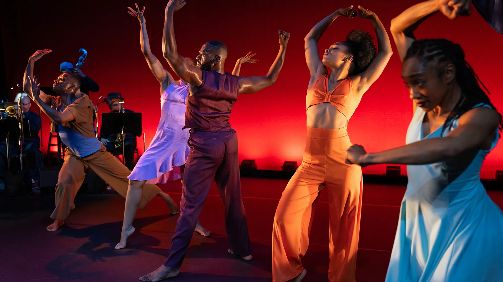 Dancers on stage strike unique poses during a performance of Dianne McIntyre's "In the Same Tongue." Their positions resonate with strength and beauty. Behind them, a live band also performs on stage.