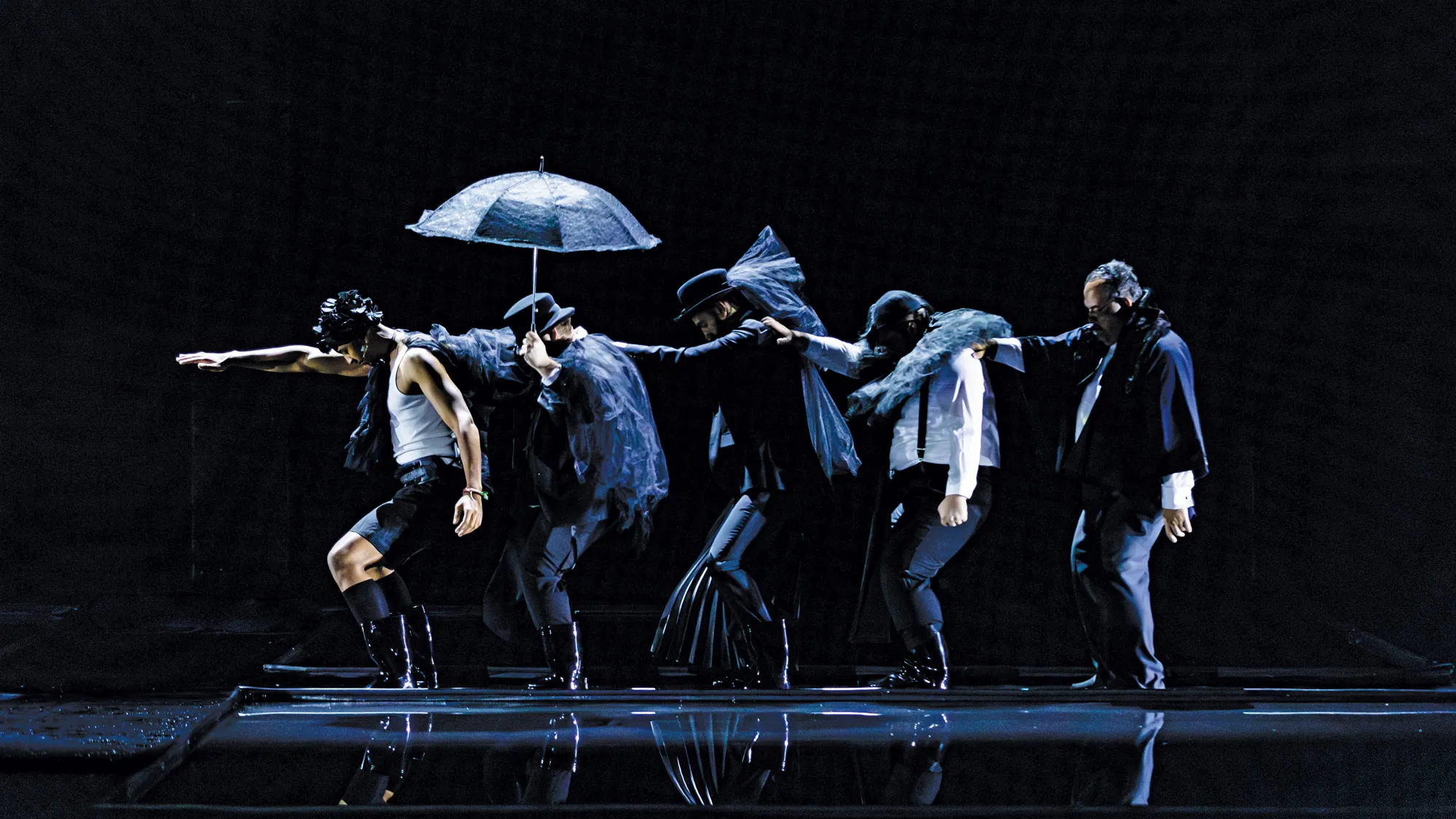 Four modernly styled actors wear black in front of a black background. Each of the five men holds the shoulder of the man in front of him. One holds an umbrella; several of the others wear clothes or boots made of water-repelling plastic. The floor seems to be wet.