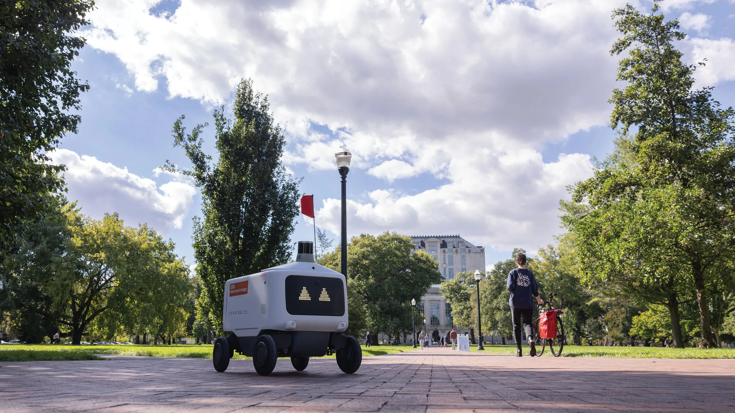 A lone food rover, which is a plastic-covered rectangle with a screen face showing triangle-shape eyes, makes its way across the oval. Thompson Library and students can be seen in the distance.