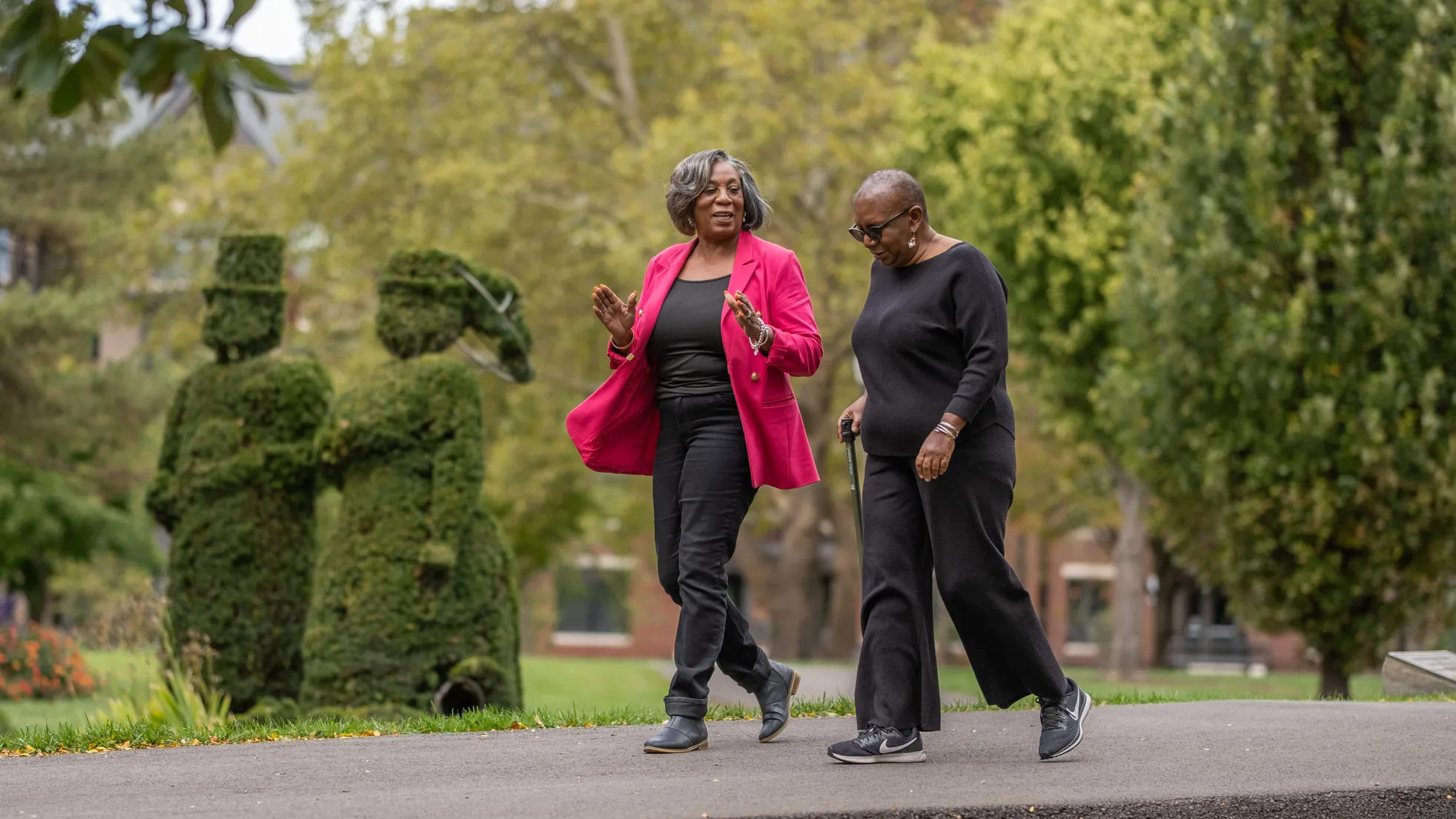 Two black ladies smile as they chat and walk through a famous park in downtown Columbus. One uses a cane and wears head-to-toe black. The other wears a bright pink suit jacket over her black outfit.