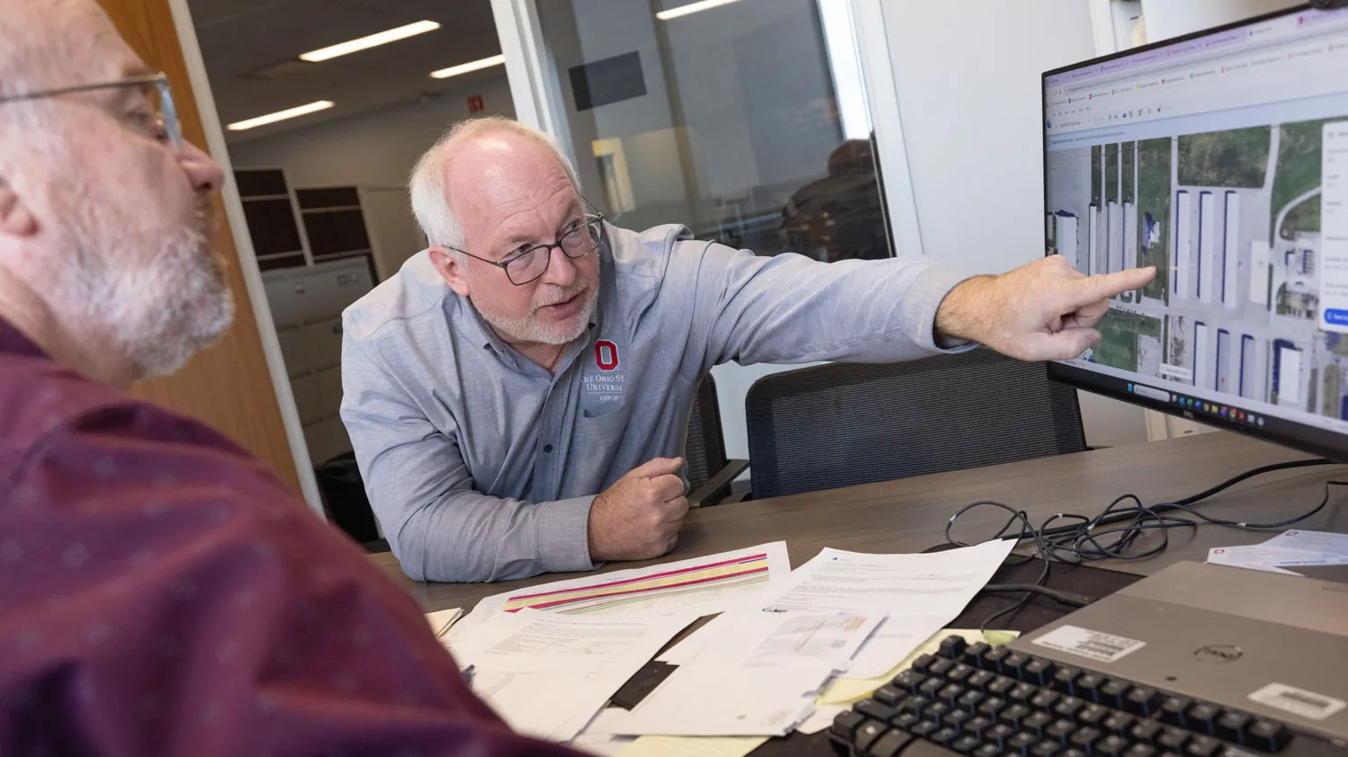 In an office at the Ohio State Airport, two men sit at a desk covered with papers and a keyboard while conferring in front of a computer monitor. Dale Gelter points toward a map or aerial image displayed on the screen, and Karl Von Hagel follows along. 