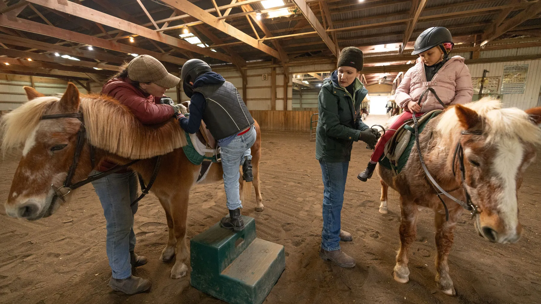 Lloyd and one of her teachers assist two riders mounting ponies inside an indoor riding arena. Lloyd adjusts one seated child while watching the teacher steady the other child, who is using a mounting block to get on their pony.