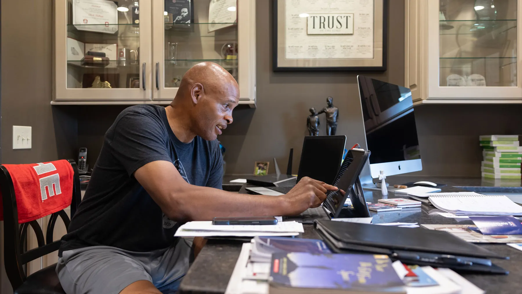 Wearing a T-shirt and gym shorts, Kellogg focuses on his iPad on a desk in his home office, surrounded by papers, notebooks, and books. On the wall behind him is a large framed picture that says “trust” along with certificates and decorative items. 