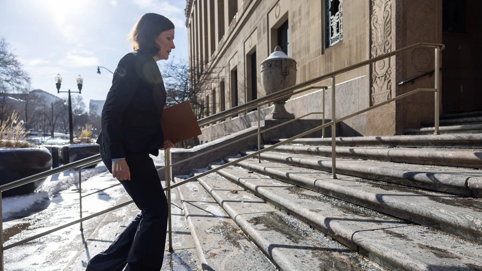 Holding a folder, Lloyd walks up a wide set of stone steps outside a courthouse, a large stone building with tall columns and railings.