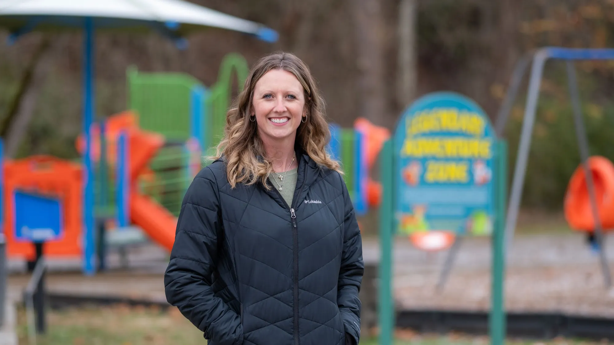A mom smiles as she poses for a photo in front of a colorful playground on a cold day