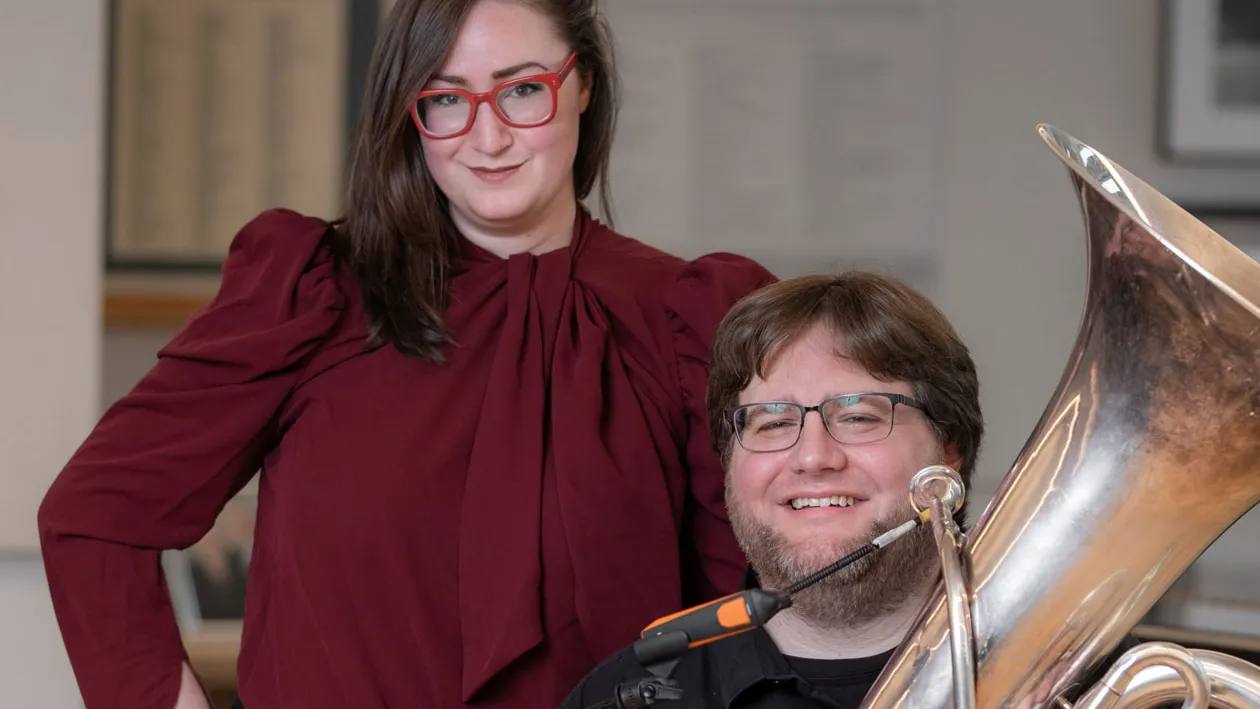 A young man smiles as he sits holding a tuba behind a microphone stand. His teacher stands close beside him with one hand on her hip. She wears bright red glasses and smiles with closed lips. She looks knowing and approving. They’re inside a music practice room.
