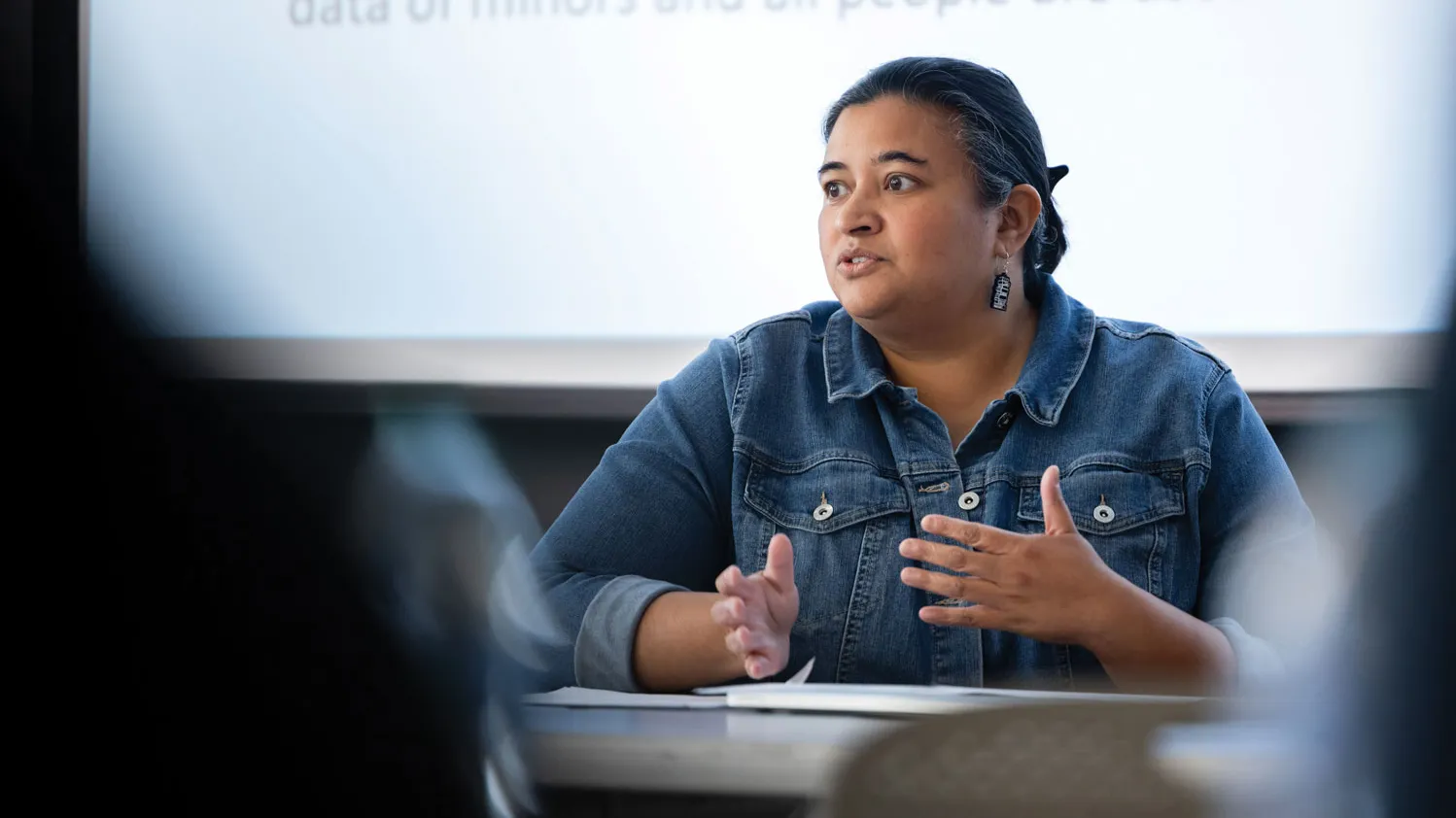As part of a panel discussion, a woman speaks in front of an audience. She gestures with her hands and wears a denim shift and hair neatly pulled back.