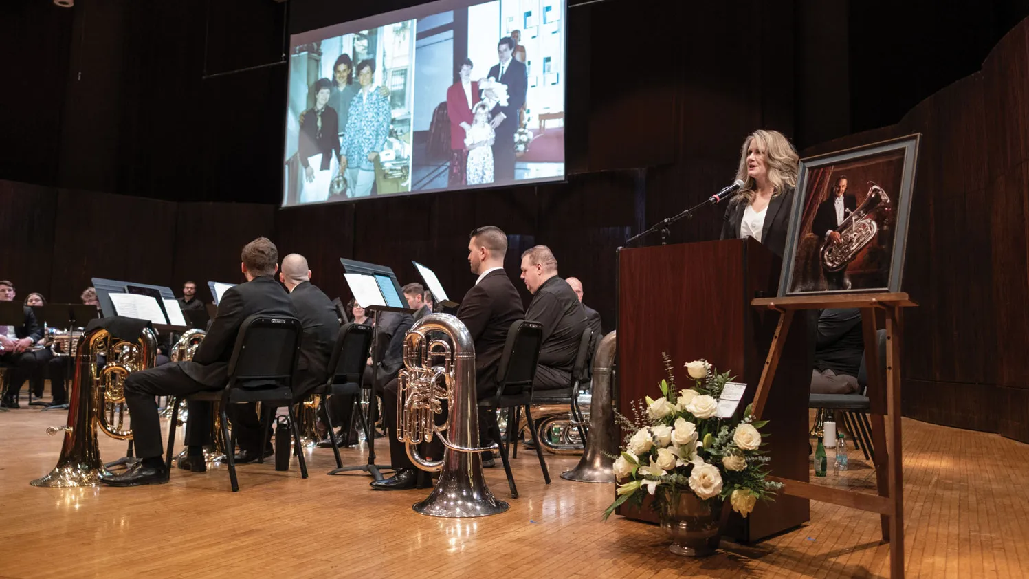 At a celebration of Jim Akins’ life, a group of musicians sit on a stage holding their instruments. To the right, his daughter speaks at a podium about her dad. His framed portrait sits on an easel nearby. Above the stage, a screen shows family photos.
