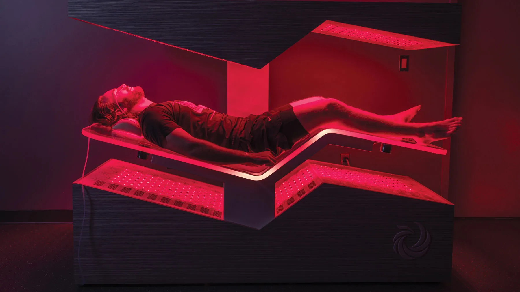 A man lies in a futuristic bedlike machine. The machine surrounds his body above and below in a structured, enclosed shape. His legs are slightly elevated and his arms rest beside his torso. Red light shines over him.