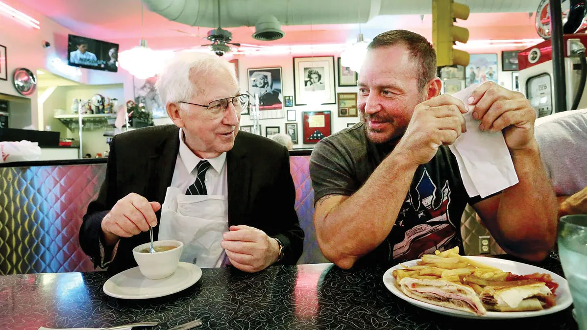 This image shows two people seated next to each other at a diner-style booth, engaged in conversation while eating. Hugh Dorrian is an older white man with white hair and a friendly, grandfatherly smile. He has a small bowl of soup. The other man is younger and wears a T-shirt and has a sandwich and fries.