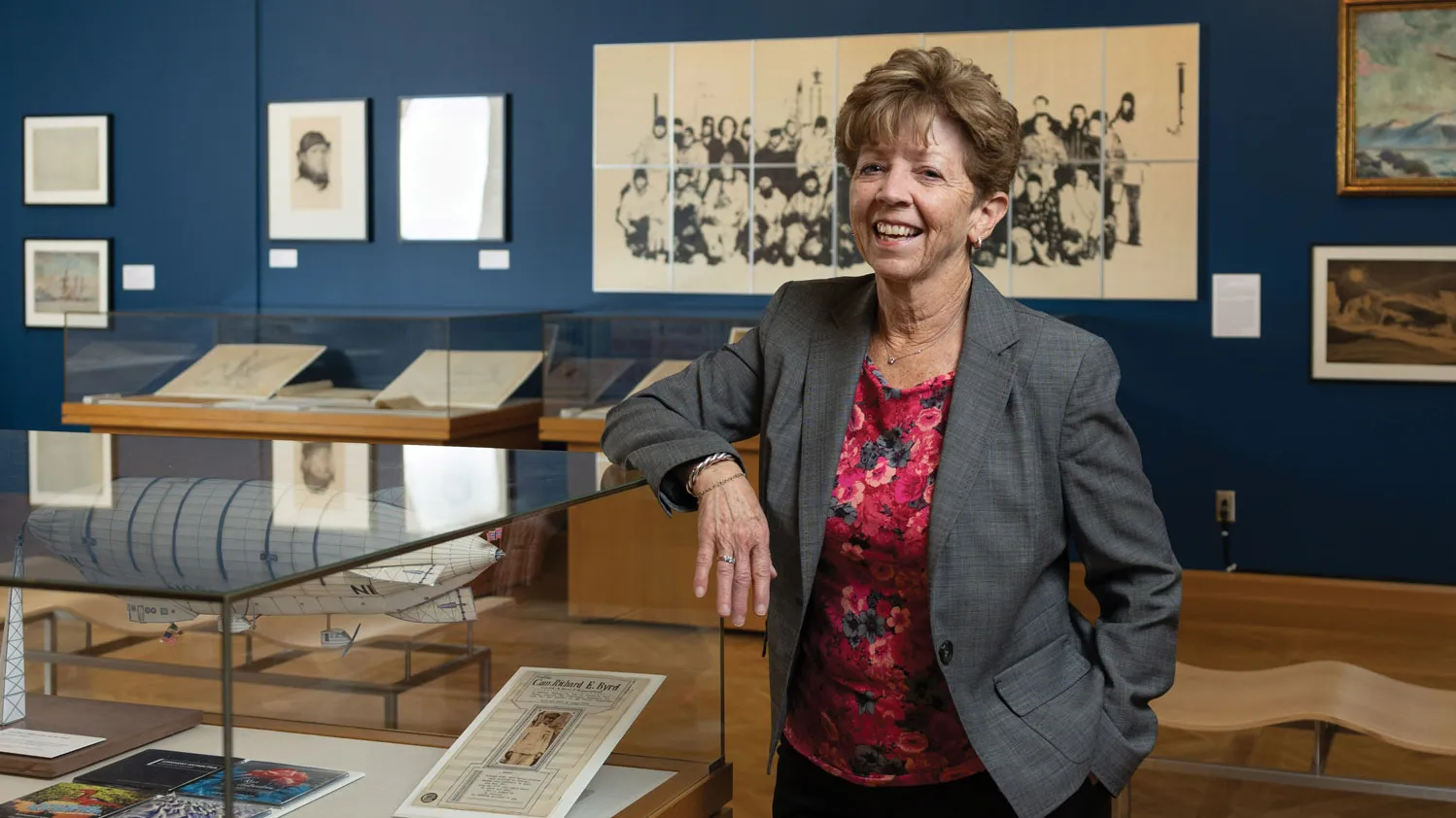 Laura Kissel, a white woman with a shorter haircut, stands in a museum exhibit she designed with polar artifacts. She’s laughing, wearing a suit coat and colorful shirt, and has an elbow propped on a glass display case. She looks like she’d be a fun co-worker.