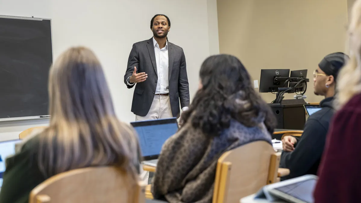 Victor St. John teaches a class. The students are seen from behind while the teacher faces the camera. He's slightly smiling as he explains his point, wearing a sport coat and button down with his long hair pulled back.