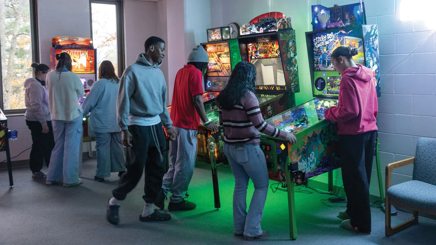 Several people stand side by side indoors, each facing a pinball machine. Colored lights shine from them onto the walls, floor and people.