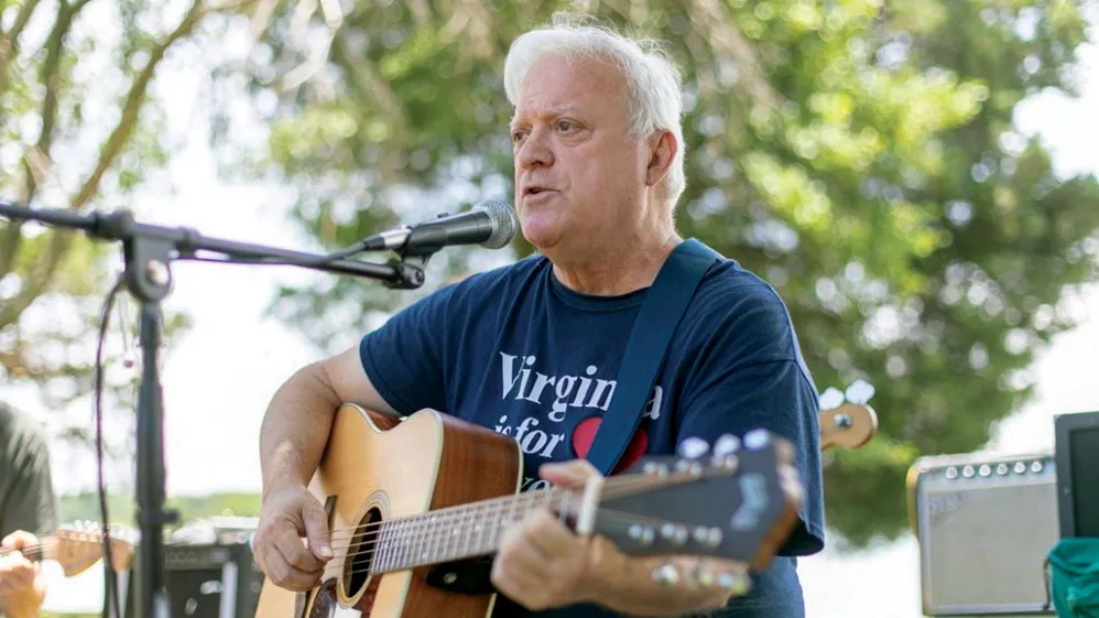 Dr. Richard Prass plays an acoustic guitar and singing into a microphone. Trees and music equipment are in the background. He’s an older white man with white hair and T-shirt that probably says “Virginia is for lovers”