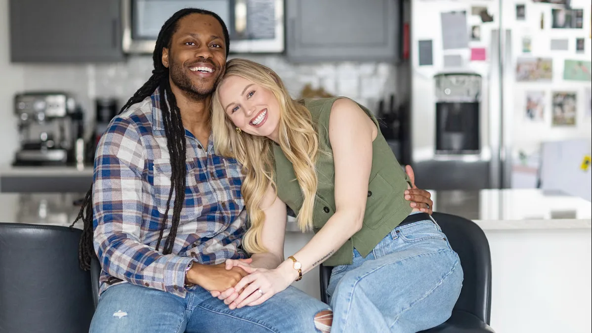 Victor St John and Christine Hamilton pose for a photo in their home. They appear happy as she leans her head on his shoulder. He's a black man with long dreds wearing a plaid button down shirt. She's a white woman with long blond hair wearing a sleeveless blouse.