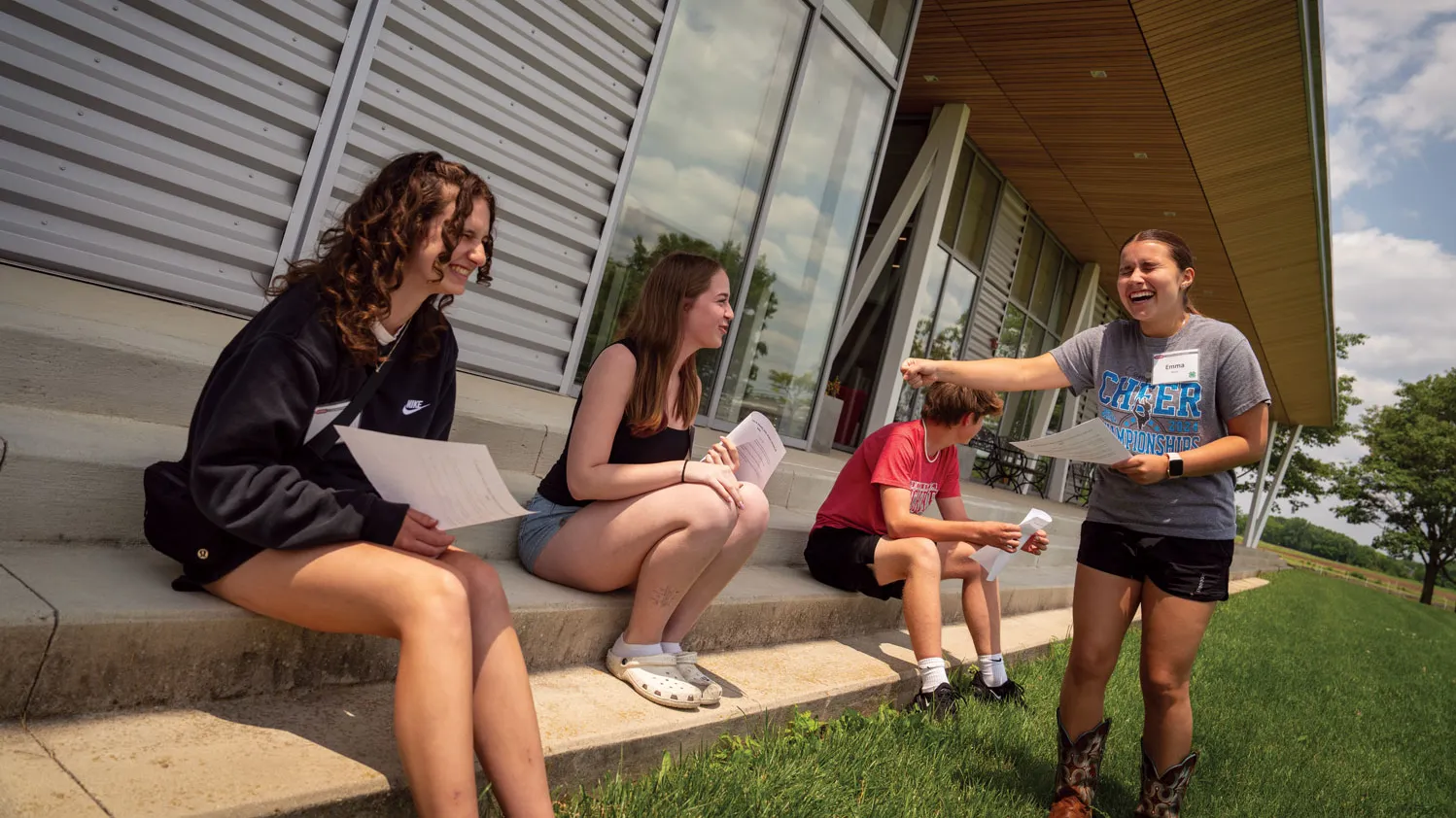 Four white teenagers laugh outside a small, modern-designed building. Three sit on a concrete sidewalk and another points and laughs. She wears cowgirl boots in addition to her shorts and T-shirt., wearing