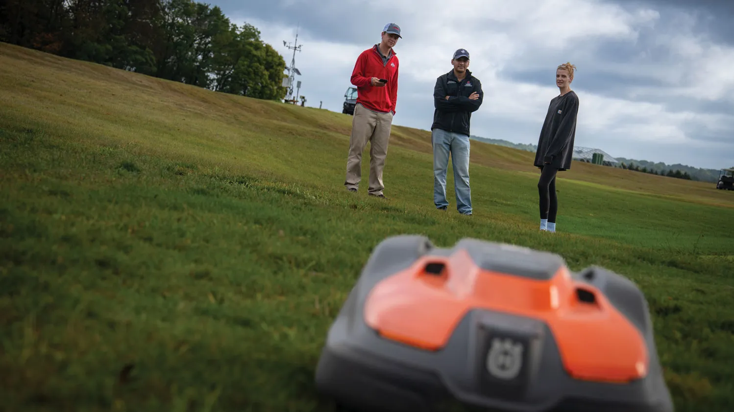 In the distance, three people stand on a lush lawn looking toward a robotic mower. That is at the front of the photo—a squarish plastic looking piece of tech that is surprisingly flat.