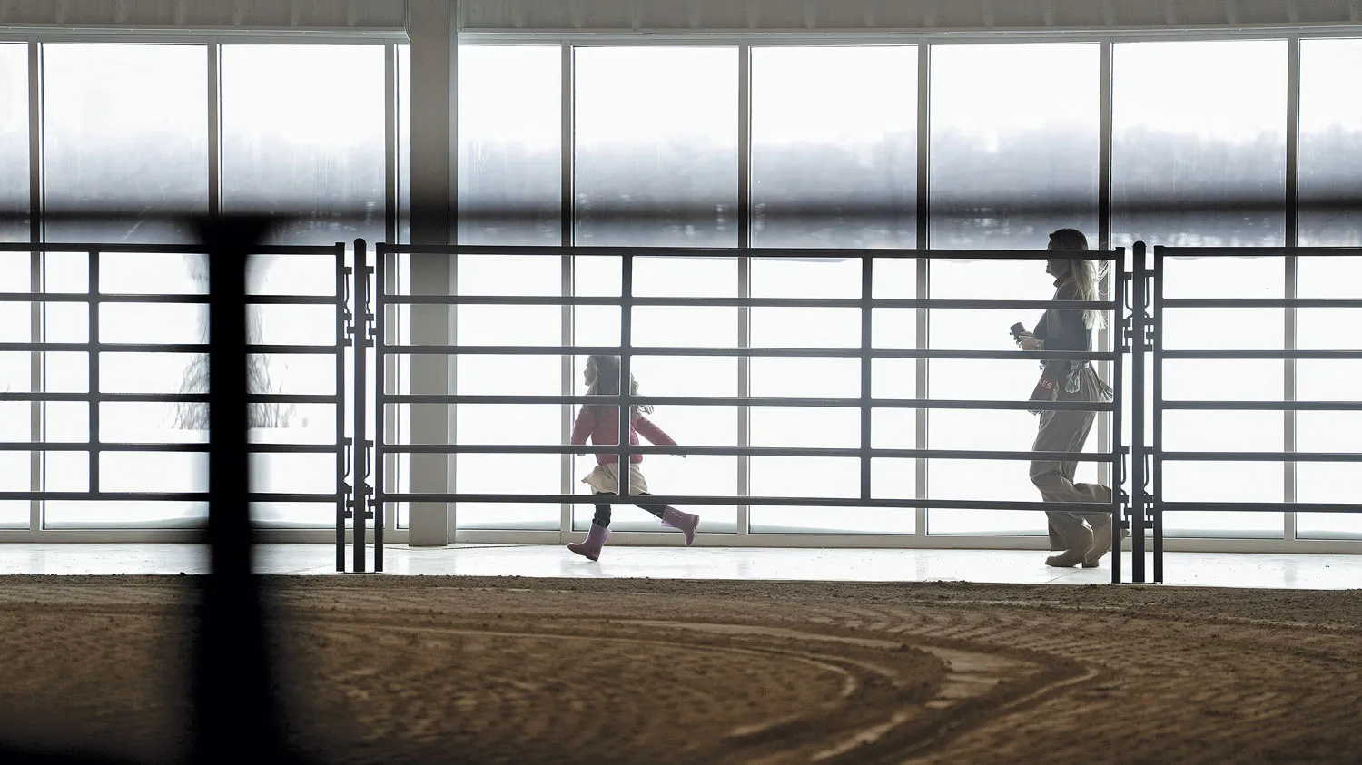 Inside a building, a girl runs between a railing taller than her head and a wall of windows. On the side of the railing closer to us is a dirt ring, such as for exercising horses. On the other side of the windows are a snowy field and, far off, a woods. More snow is falling. The girl’s mother trails her.