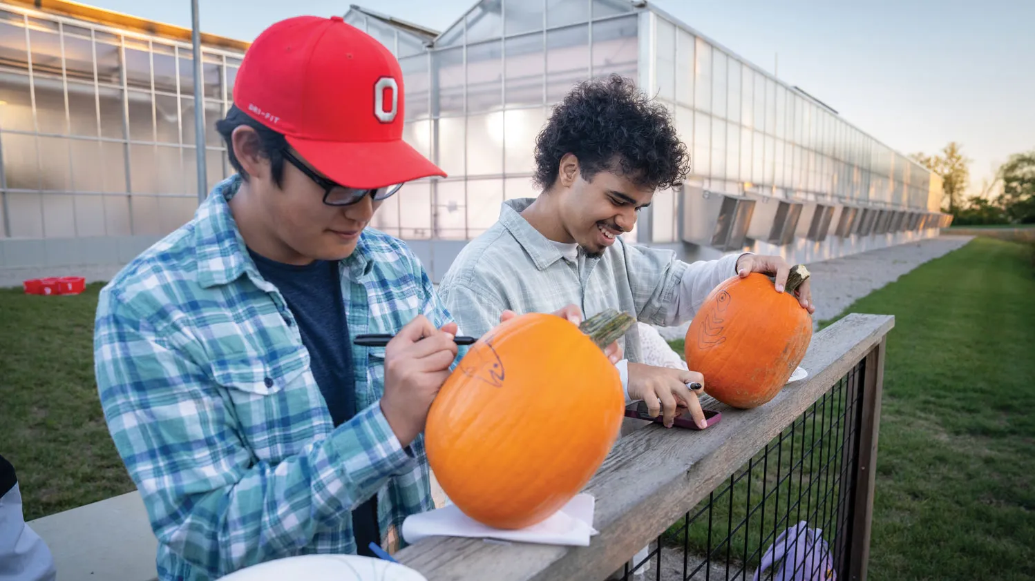 Two young men draw with markers on pumpkins that are taller than round. One young man is focusing on his drawing; the other laughs as he checks his phone--presumably a design inspiration.