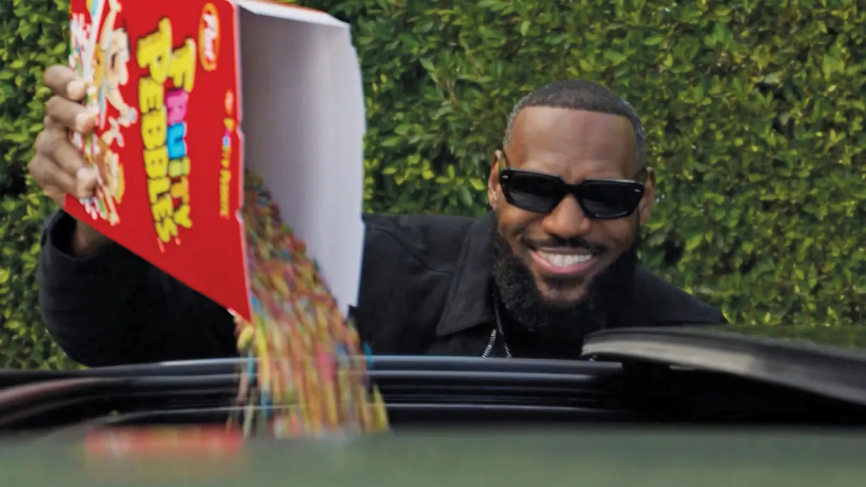 Lebron grins as he pours a box of Fruity Pebbles through the sunroof of a car. The basketball icon is a black man with beard longer than the hair on top of his head and wearing sunglasses.
