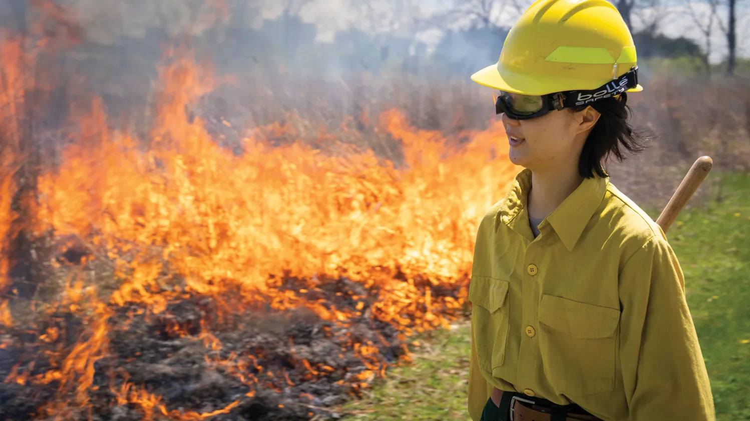 A young Asian woman wears safety goggles, a yellow hard hat and matching work shirt as she watches a fire burn over a patch of dark ground. Alongside is green grass.