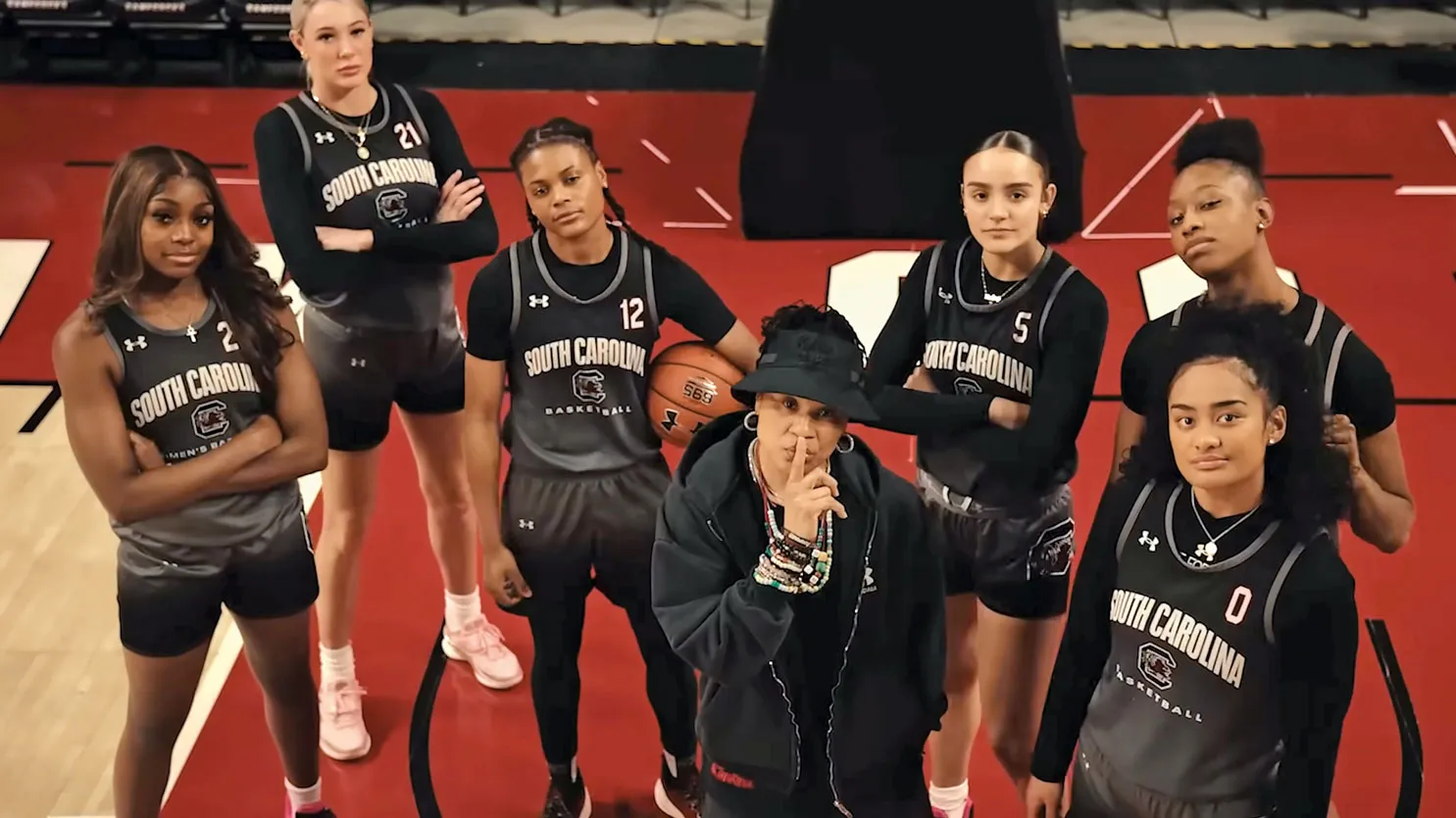 Standing on a basketball court, a college coach makes a "shh" gesture in front of her lips, while six of her basketball players stand around her posing to look tough. 