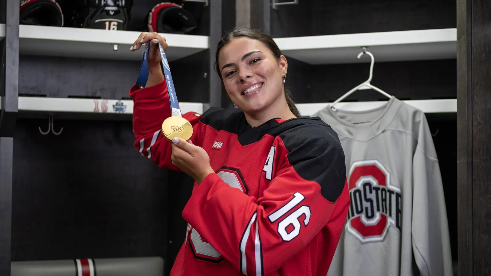 Joy Dunne smiles as she proudly shows off her gold medal in the locker room for Ohio State women's hockey. She wears her team uniform. 