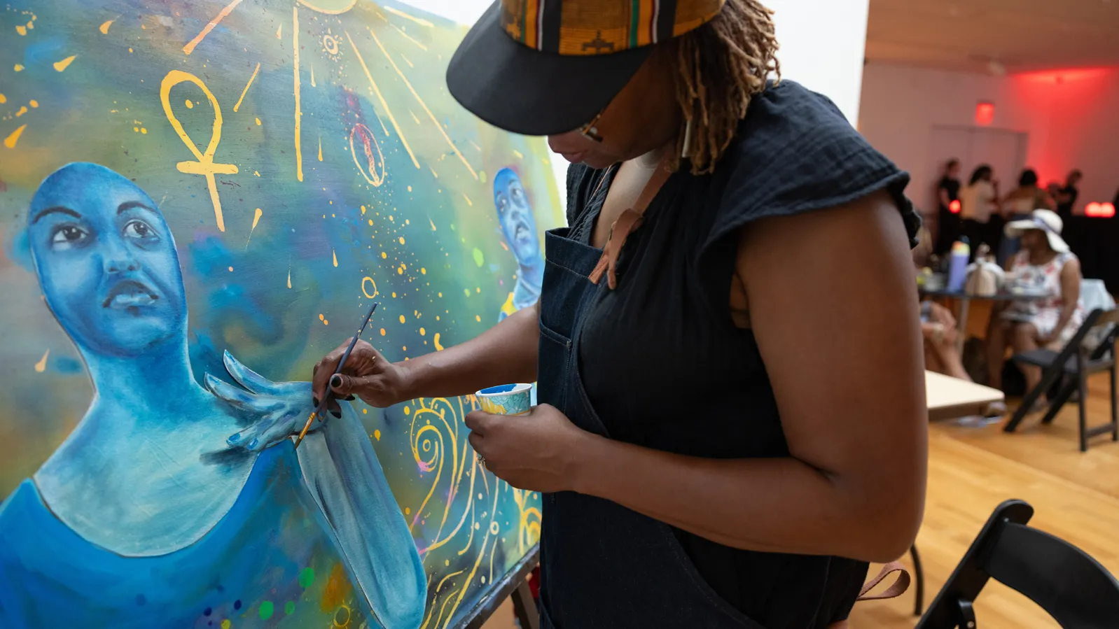 Sunami paints, wearing an apron and ballcap, at an event. In the background, there are people talking and sitting at tables. Her painting is in shades of blue, green and gold, and it focuses on a woman