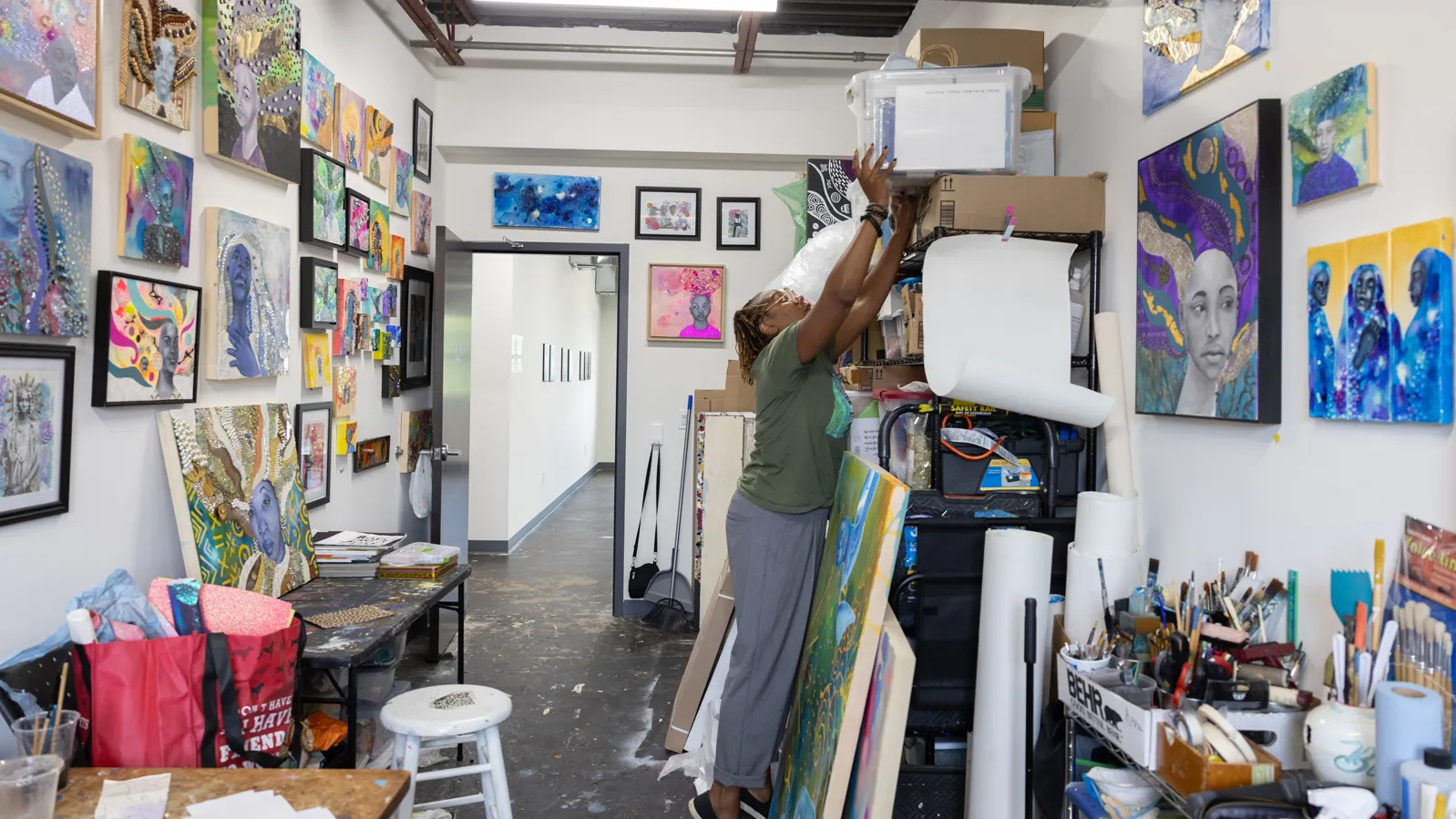 Sunami reaches higher than she is tall for a plastic bin on top of shelves and other stacks of supplies. She's in her studio which is full of interesting paintings and tools 