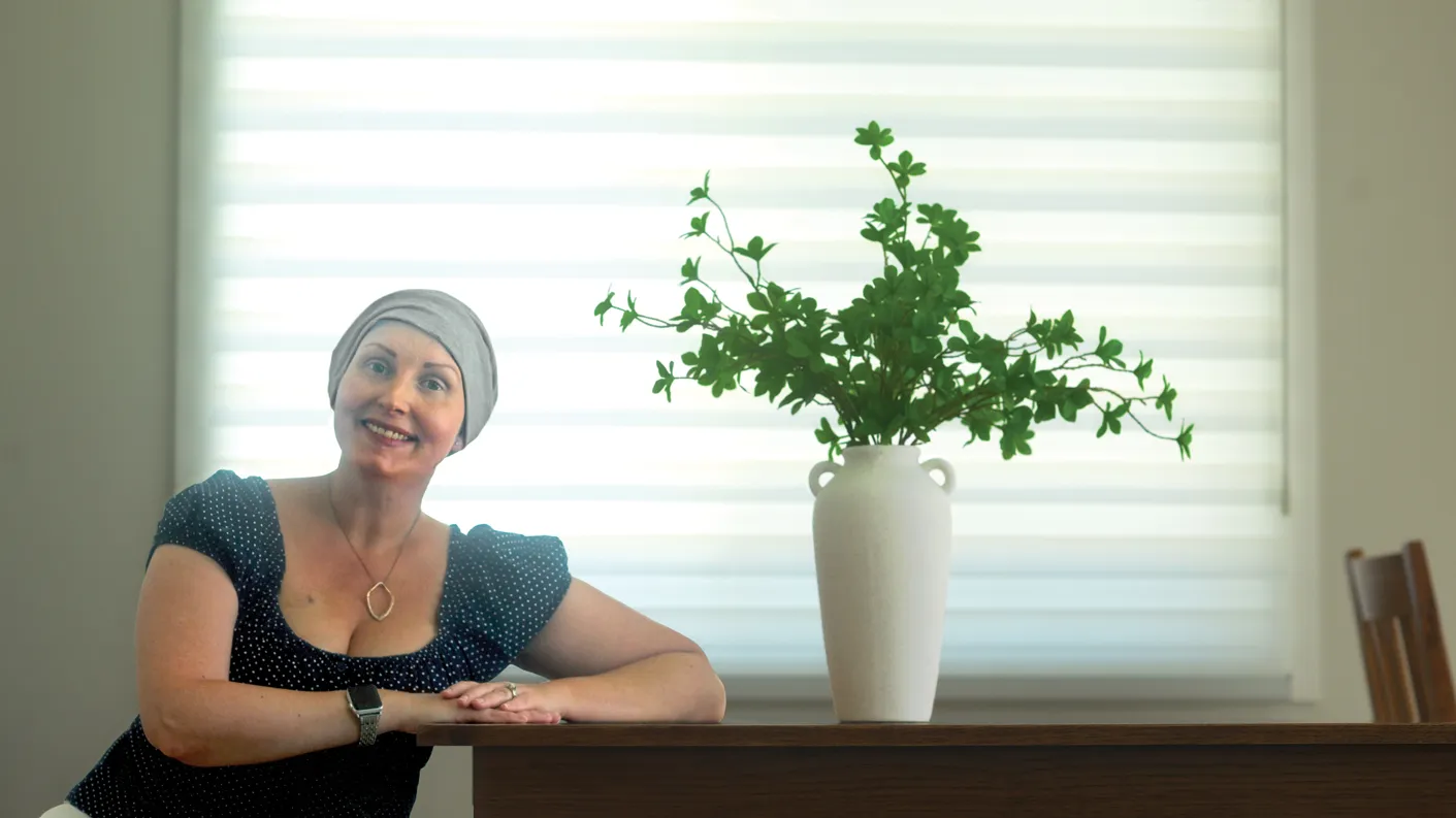 Stephanie Balm smiles as she sits at a table with a lush looking plant in a vase.