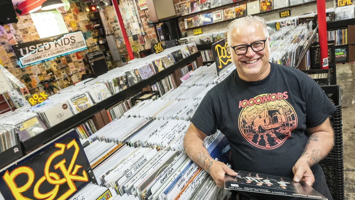 Used Kids owner Greg Hall laughs as he is photographed in the record store, wearing thick-framed glasses and an artful T-shirt for the hard rock band 1000 Mods.