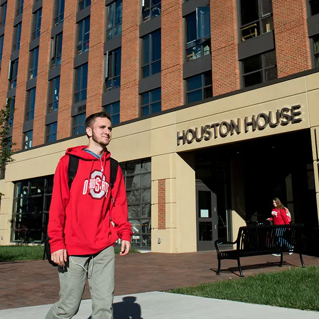 A young man in a red Ohio State sweatshirt is walking by a brick building named "Houston House". 